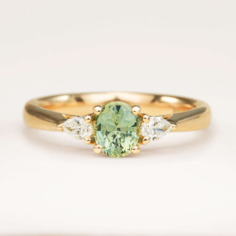 An oval-cut teal sapphire just under 1ct and pear-cut diamond three-stone engagement ring set in yellow gold, displayed on a clean white background.