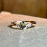 An oval-cut greeny-teal sapphire and diamond three-stone engagement ring, displayed on a book.