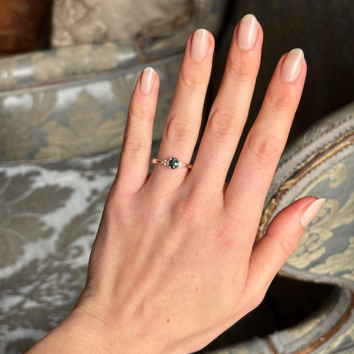 Teal sapphire and diamond three-stone engagement ring with oval-cut centre stone, displayed on a hand, emphasising its sparkling diamonds and rich colour from a distance.