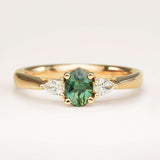 Oval-cut teal sapphire and diamond three-stone engagement ring resting on a book, showcasing the centre sapphire and diamond accents.