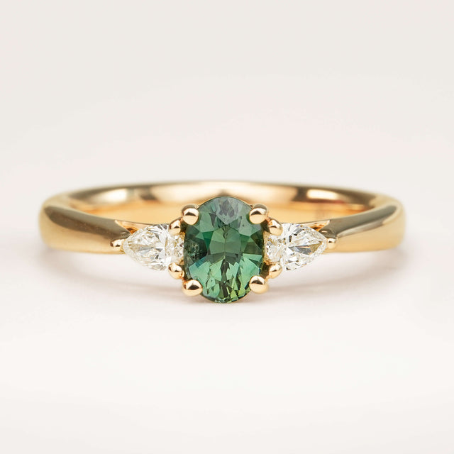 Oval-cut teal sapphire and diamond three-stone engagement ring resting on a book, showcasing the centre sapphire and diamond accents.