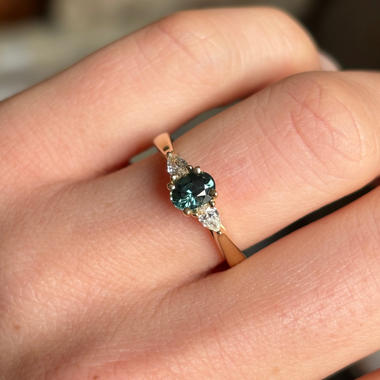 Oval-cut teal sapphire and diamond three-stone engagement ring worn on the engagement ring finger, showing its elegance and fit.