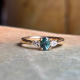 Oval-cut teal sapphire and diamond three-stone engagement ring resting on a book, showcasing the centre sapphire and diamond accents.
