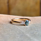Close-up of an oval-cut teal sapphire centre with diamond accents in a three-stone engagement ring, displayed on a old book background to emphasise colour and craftsmanship.