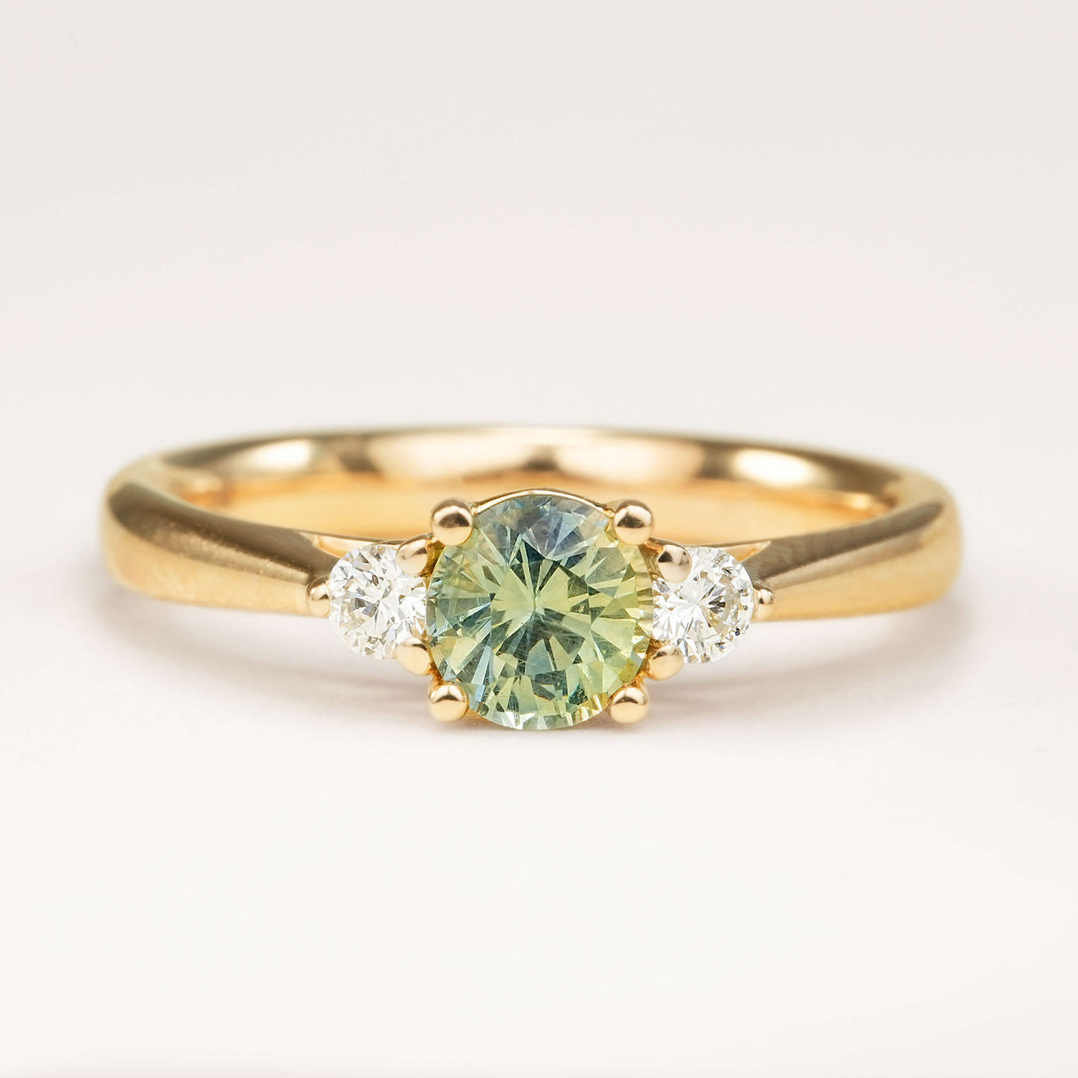 Round-cut teal sapphire and diamond three-stone engagement ring displayed on a professional off-white background, highlighting its true colour and classic design.
