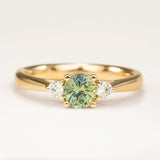 Round-cut teal sapphire and diamond three-stone engagement ring displayed on a professional off-white background, highlighting its true colour and classic design.