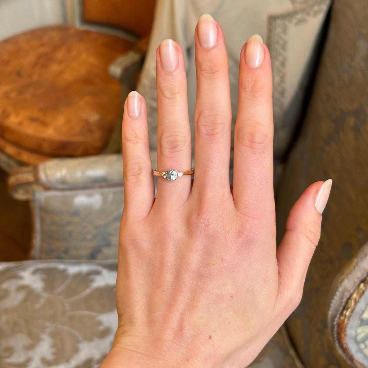 Round-cut teal sapphire and diamond three-stone engagement ring worn on the hand, emphasising the sapphire’s size and diamond accents.