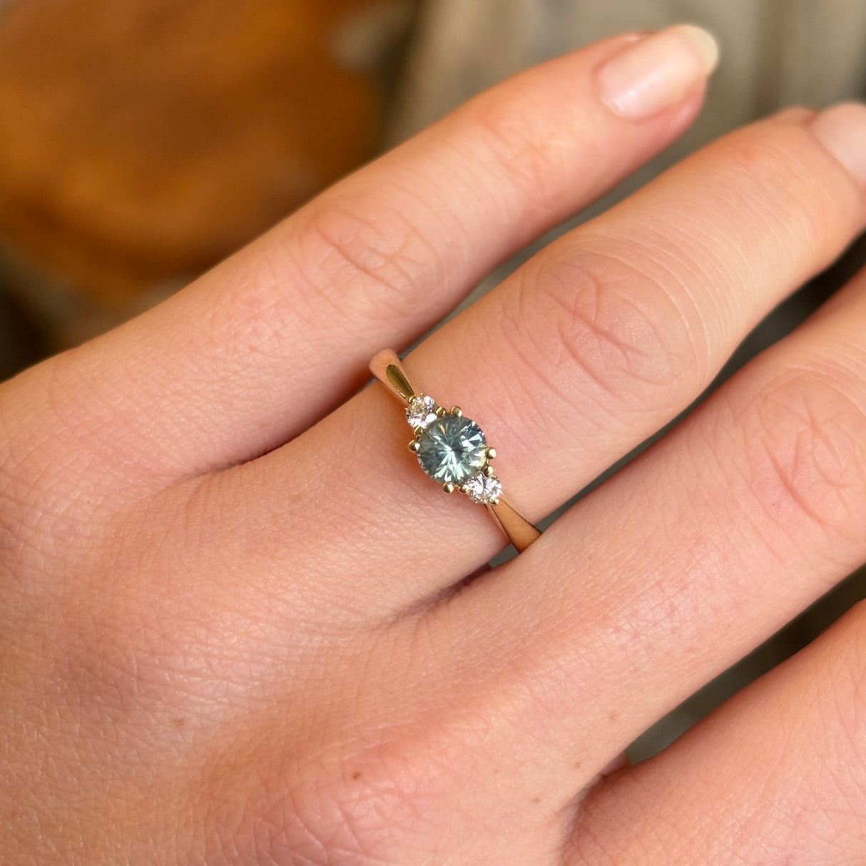 Round-cut teal sapphire and diamond three-stone engagement ring worn on the hand, illustrating its size and proportion.