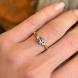 Round-cut teal sapphire and diamond three-stone engagement ring worn on the hand, illustrating its size and proportion.