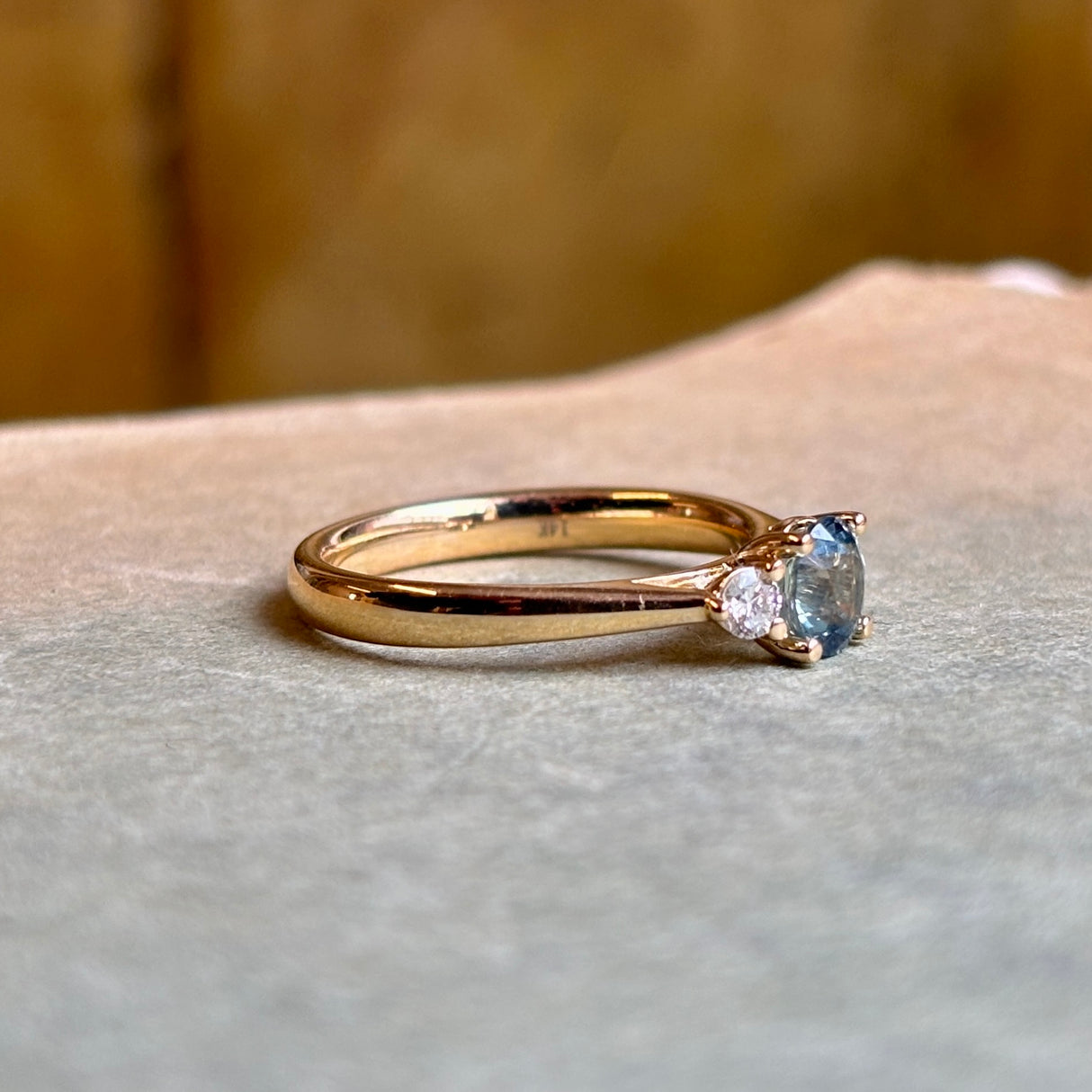 Round-cut teal sapphire and diamond three-stone engagement ring displayed on a book, showing the design and width of the band.