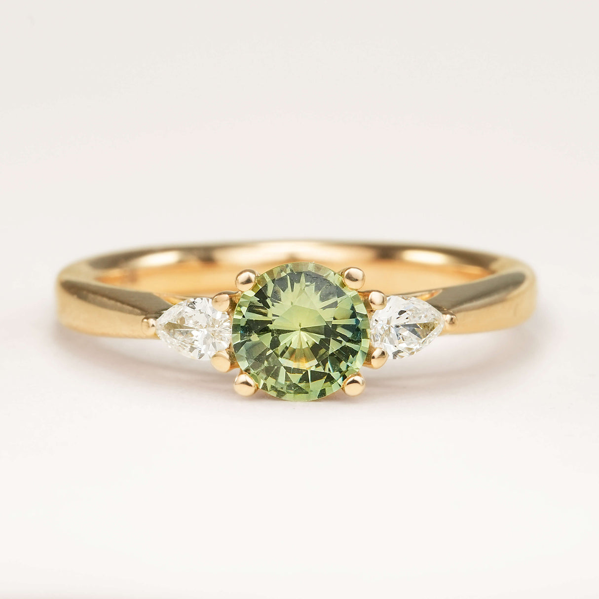 Round bright teal sapphire and pear-cut diamond three-stone engagement ring displayed on an off-white background, highlighting its colour and design.