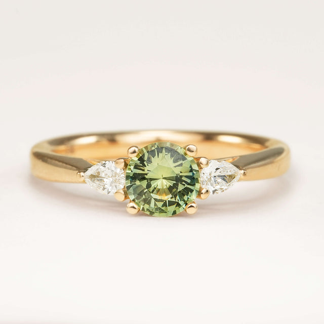 Round bright teal sapphire and pear-cut diamond three-stone engagement ring displayed on an off-white background, highlighting its colour and design.
