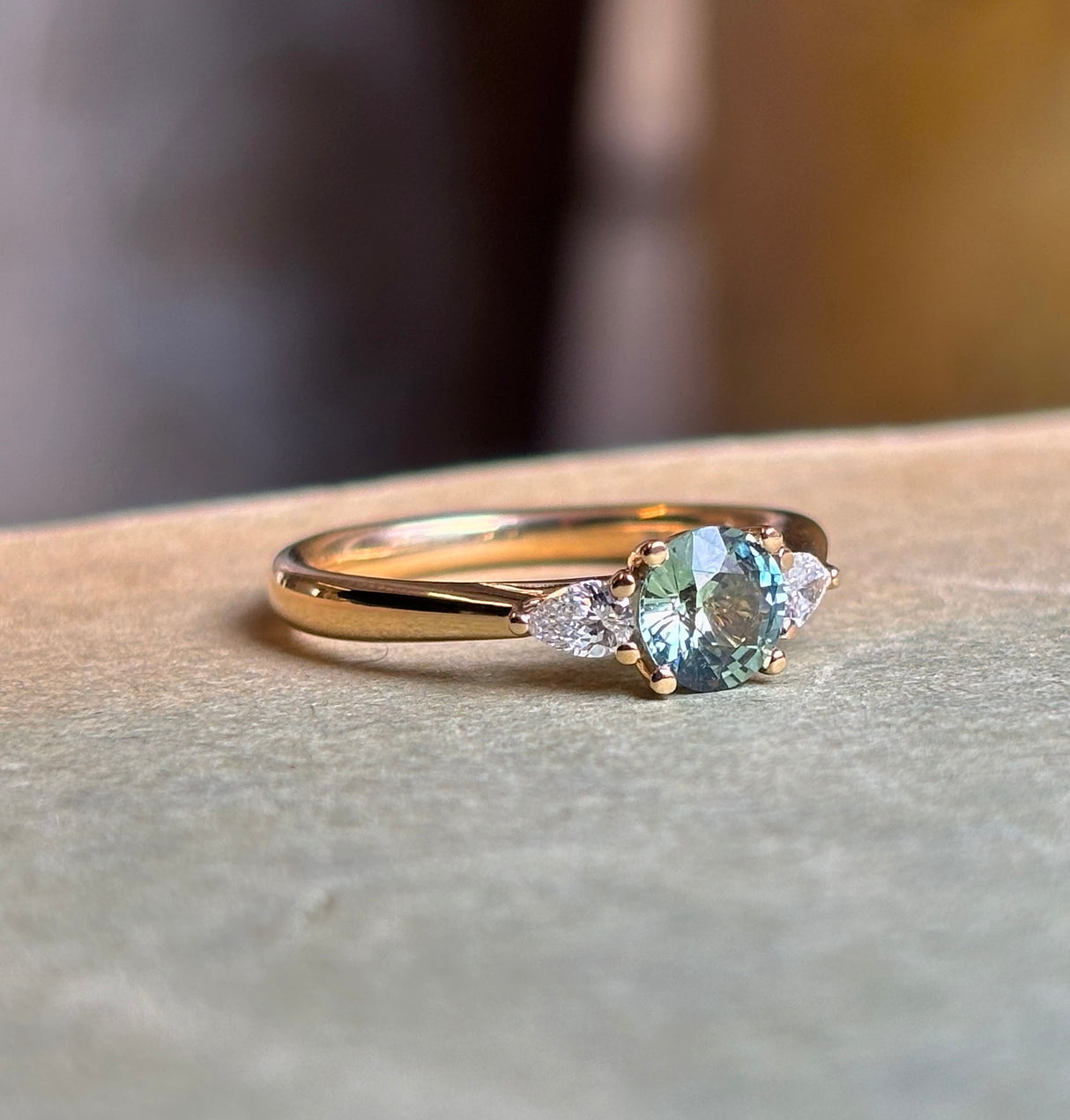 Round teal sapphire and pear-cut diamond three-stone engagement ring resting on a book, showcasing the sapphire centre and pear-cut diamond accents.