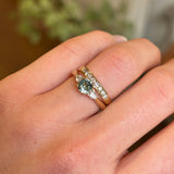 Round Teal Sapphire and Pear-Cut Diamond Three-Stone Engagement Ring Worn on Engagement Ring Finger next to a wedding band.