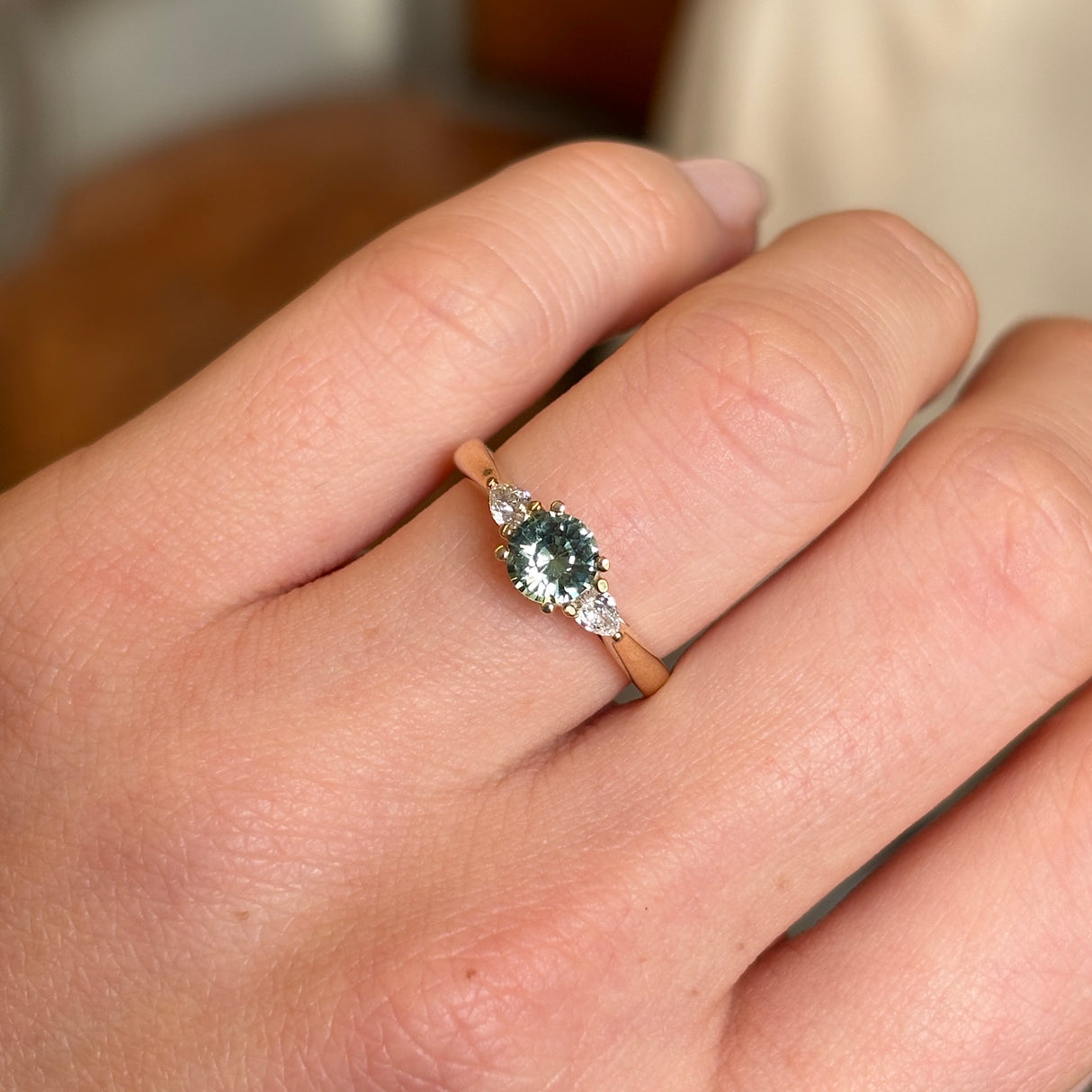 A bright central round teal sapphire and pear-cut diamond three-stone engagement ring worn on the hand, highlighting the sapphire’s colour and pear-cut diamonds.