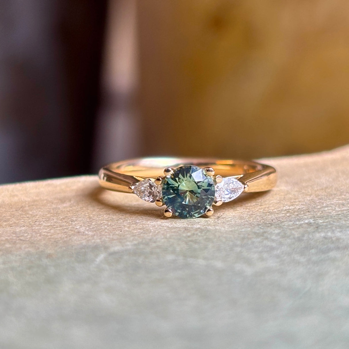 Round teal sapphire and pear-cut diamond three-stone engagement ring resting on a book, showcasing the sapphire centre and pear-cut diamond accents.
