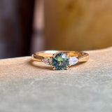 Round teal sapphire and pear-cut diamond three-stone engagement ring resting on a book, showcasing the sapphire centre and pear-cut diamond accents.