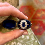 Hand holding a sapphire and diamond cluster ring in a blue ringbox