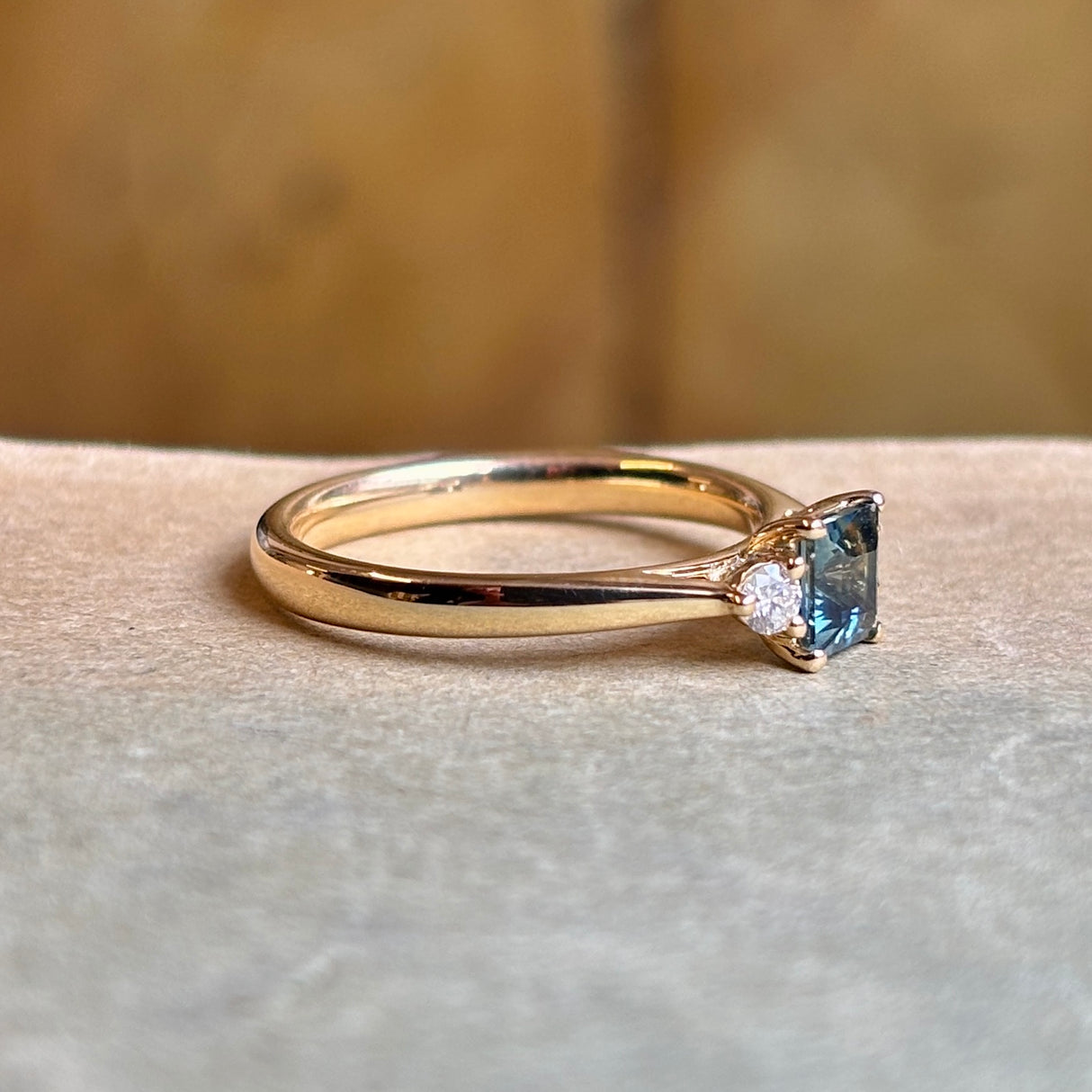 Side profile view of a square-cut teal sapphire and diamond three-stone engagement ring.