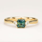 A square-cut teal sapphire and diamond three-stone engagement ring, displayed on a clean white background, highlighting its colour, elegant design and classic shape.