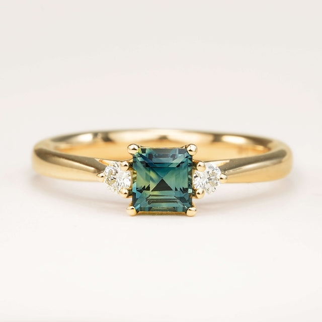 A square-cut teal sapphire and diamond three-stone engagement ring, displayed on a clean white background, highlighting its colour, elegant design and classic shape.