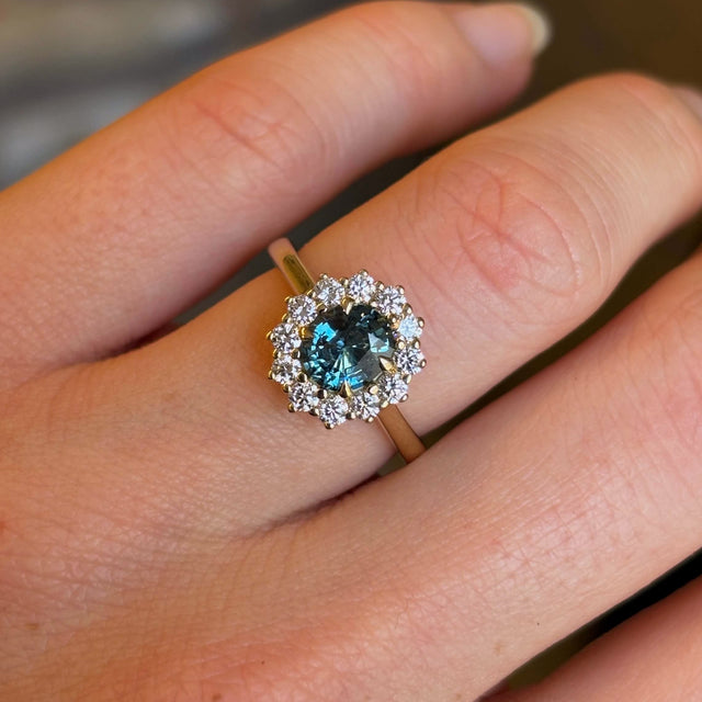 Teal sapphire and diamond cluster engagement ring captured up close on hand. 