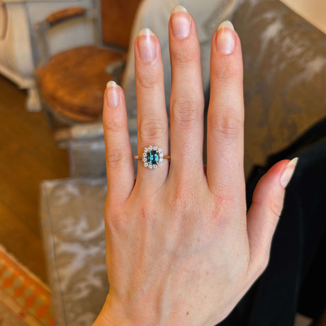 Teal sapphire and diamond cluster engagement ring shown on ring finger of left hand