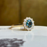 Teal sapphire and diamond yellow gold cluster engagement ring angled on light background. 