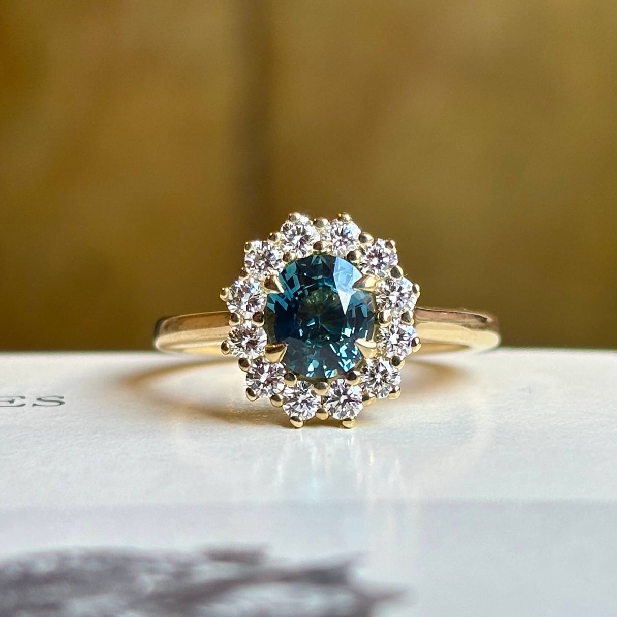 Teal sapphire and diamond yellow gold cluster engagement ring on light background. 