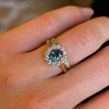 Teal sapphire and diamond cluster engagement ring worn with wedding band on hand close up 
