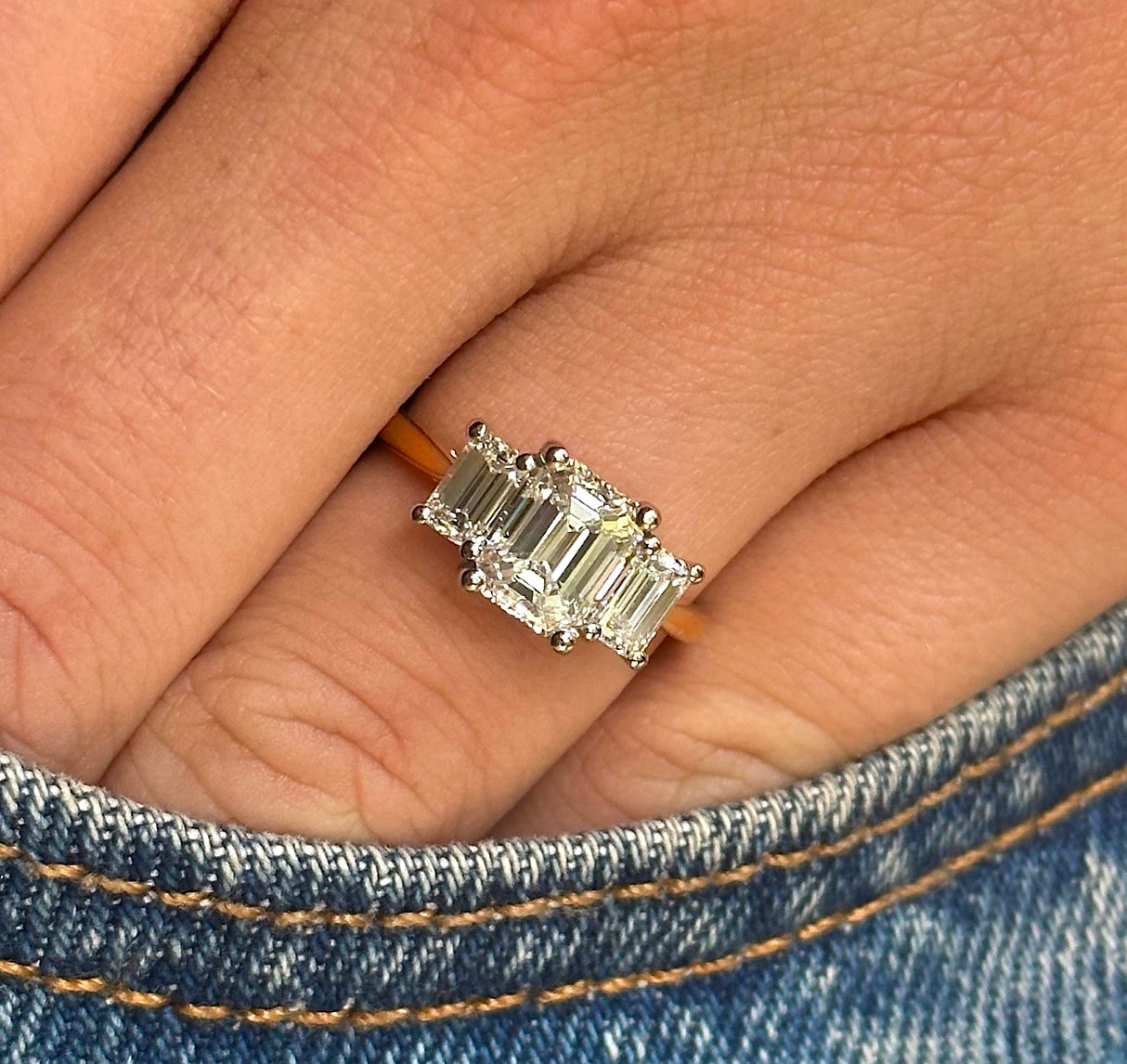 Three stone emerald-cut diamond ring, worn on middle finger photographed on hand inside denim jean pocket