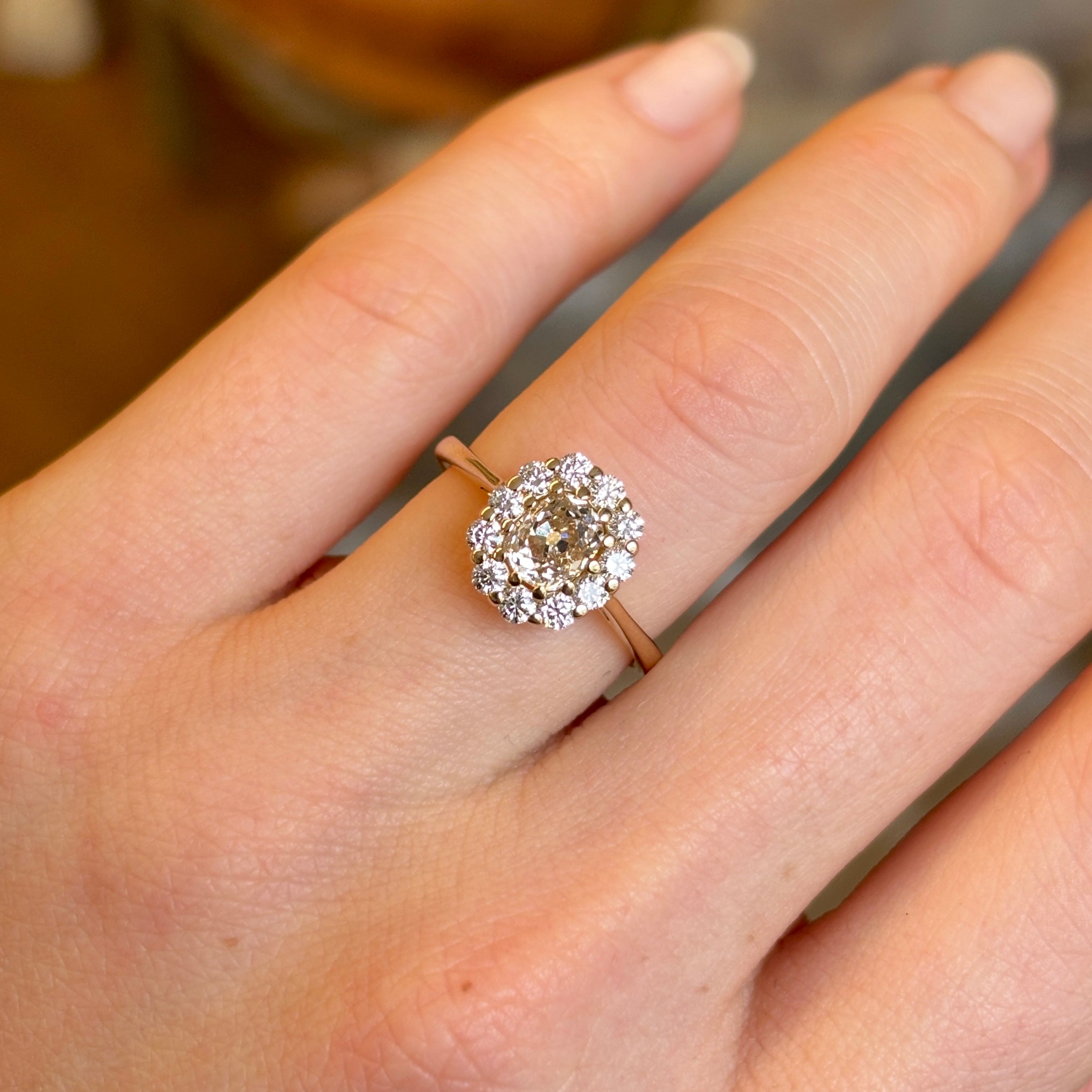 View of a yellow-brown tinted old mine-cut diamond cluster engagement ring worn on the engagement ring finger, captured in natural light.