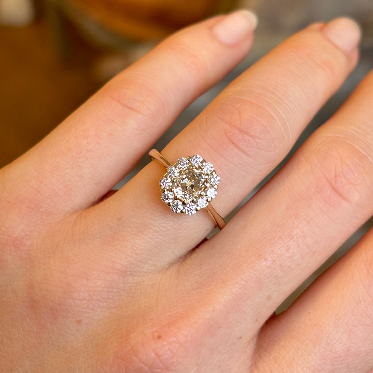 View of a yellow-brown tinted old mine-cut diamond cluster engagement ring worn on the engagement ring finger, captured in natural light.