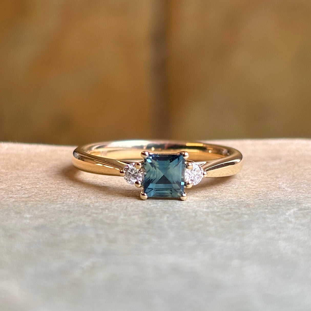 A square-cut teal sapphire and round-cut diamond three-stone engagement yellow gold ring resting on an antique book.