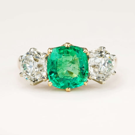 Vintage 1940s cushion-cut emerald and old-cut diamond three-stone engagement ring displayed on an off-white background, highlighting the emerald’s rich green colour and diamond sparkle.