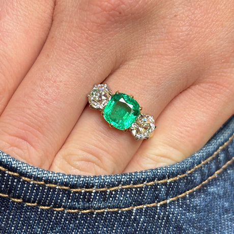 Vintage 1940s cushion-cut emerald and old-cut diamond three-stone engagement ring worn on the hand, with the hand inside a jean pocket to show scale and everyday wear.