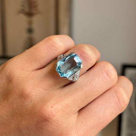 Antique Art Deco aquamarine and diamond cocktail ring in platinum shown on hand, highlighting the vibrant aquamarine centre stone and sparkling diamond surround against a softly blurred indoor backdrop.