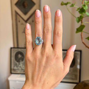Antique Art Deco aquamarine and diamond cocktail ring in platinum worn on hand, displaying a large blue aquamarine and diamond cluster against a warm vintage-inspired interior setting.