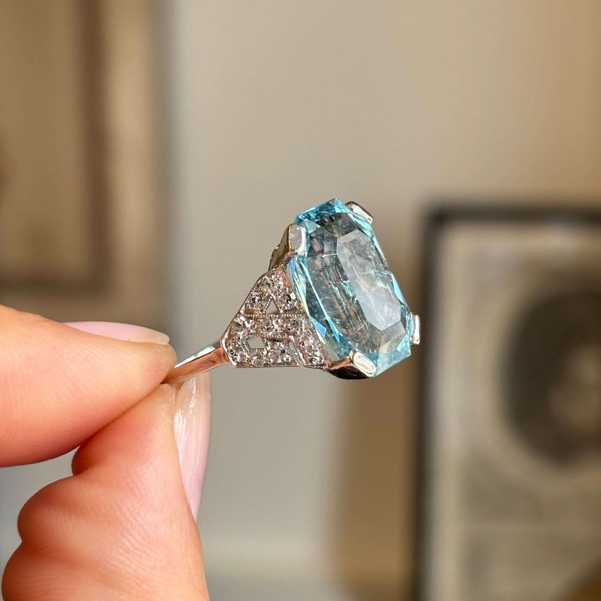 Side profile of an antique Art Deco aquamarine and diamond cocktail ring in platinum, held by hand with a vintage interior backdrop, highlighting the elevated setting and vivid blue aquamarine.