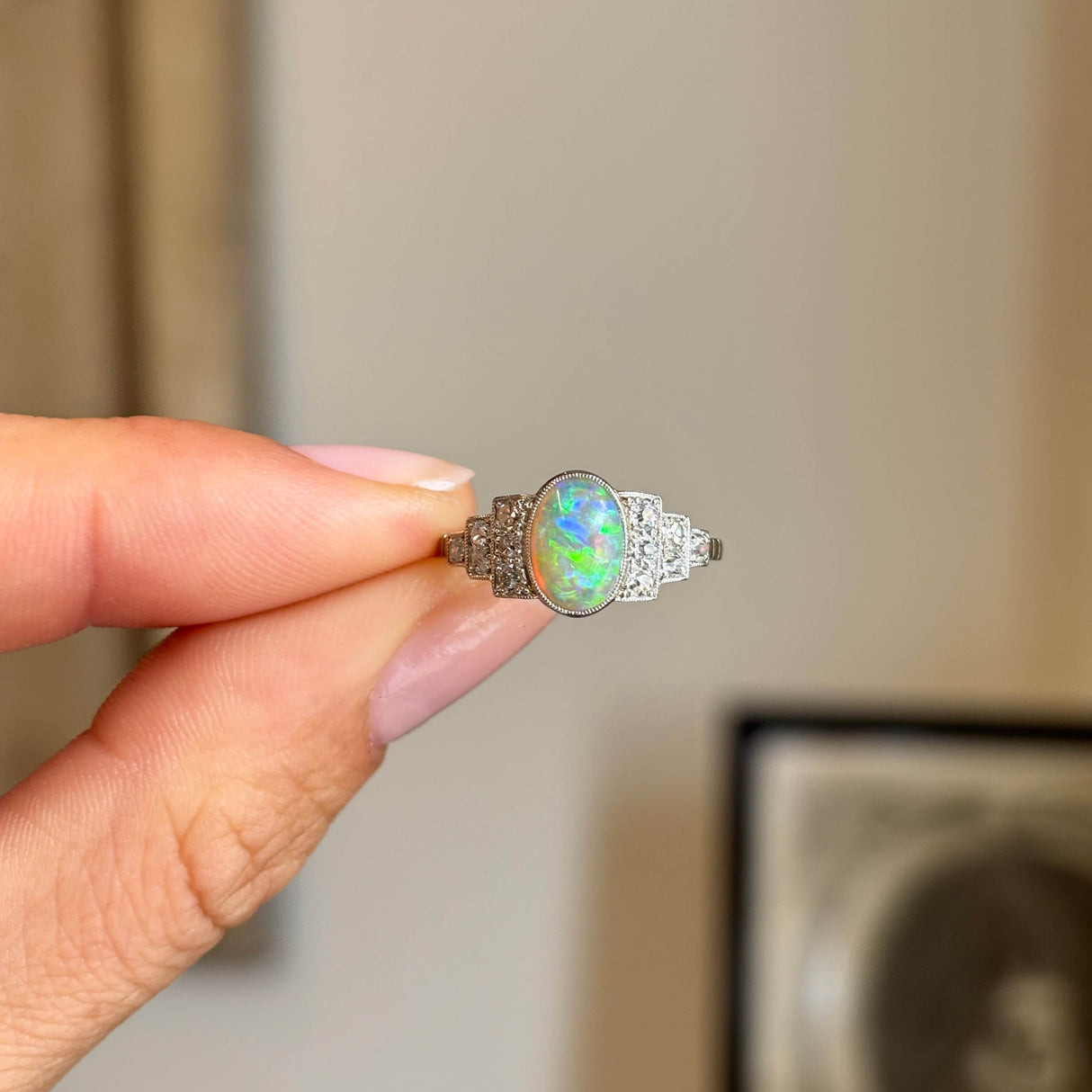 Antique Art Deco opal and diamond cluster ring held between fingers, featuring a luminous oval opal flanked by sparkling old-cut diamonds.