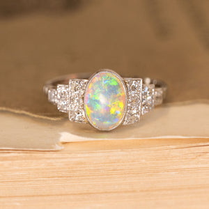 Antique Art Deco opal and diamond cluster ring featuring an oval opal displaying vivid flashes of green, blue and orange, framed by old-cut diamonds in a geometric setting.