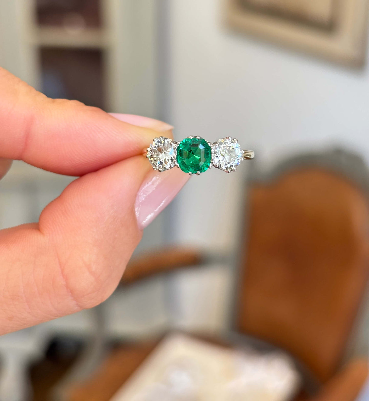 Antique Edwardian emerald and diamond three-stone engagement ring featuring an emerald centre stone flanked by two diamonds, held by hand.