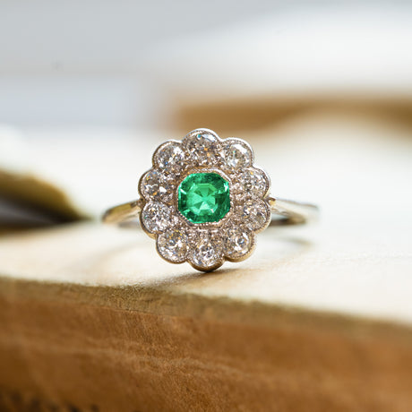 Antique Edwardian emerald and old-mine cut diamond cluster ring displayed on antique book pages with green emerald centre stone.