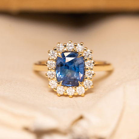 Antique-inspired blue cushion-cut sapphire and diamond cluster engagement ring featuring a richly coloured cushion-cut blue sapphire surrounded by brilliant-cut diamonds in yellow gold.