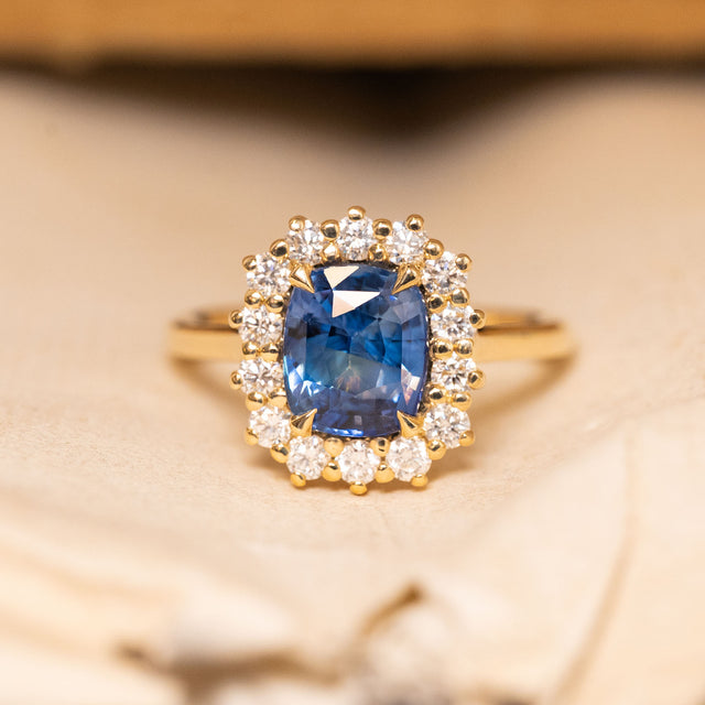 Antique-inspired blue cushion-cut sapphire and diamond cluster engagement ring featuring a richly coloured cushion-cut blue sapphire surrounded by brilliant-cut diamonds in yellow gold.