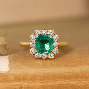 Antique-inspired emerald and diamond cluster engagement ring featuring an emerald-cut emerald surrounded by brilliant-cut diamonds in yellow gold.