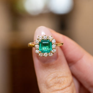 Antique-inspired emerald and diamond cluster engagement ring featuring an emerald-cut emerald surrounded by brilliant-cut diamonds in yellow gold.
