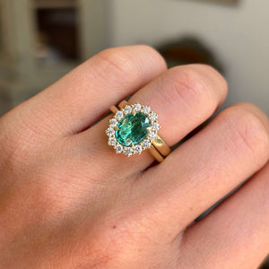 Antique-inspired yellow gold cluster ring featuring an oval-cut emerald surrounded by brilliant diamonds, photographed on hand against a softly lit vintage interior backdrop.