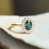 Green sapphire and diamond cluster engagement ring with oval-cut sapphire and gold band.