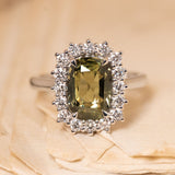 Antique-inspired green sapphire and diamond cluster engagement ring featuring a cushion-cut green sapphire surrounded by a halo of brilliant-cut diamonds in white gold.
